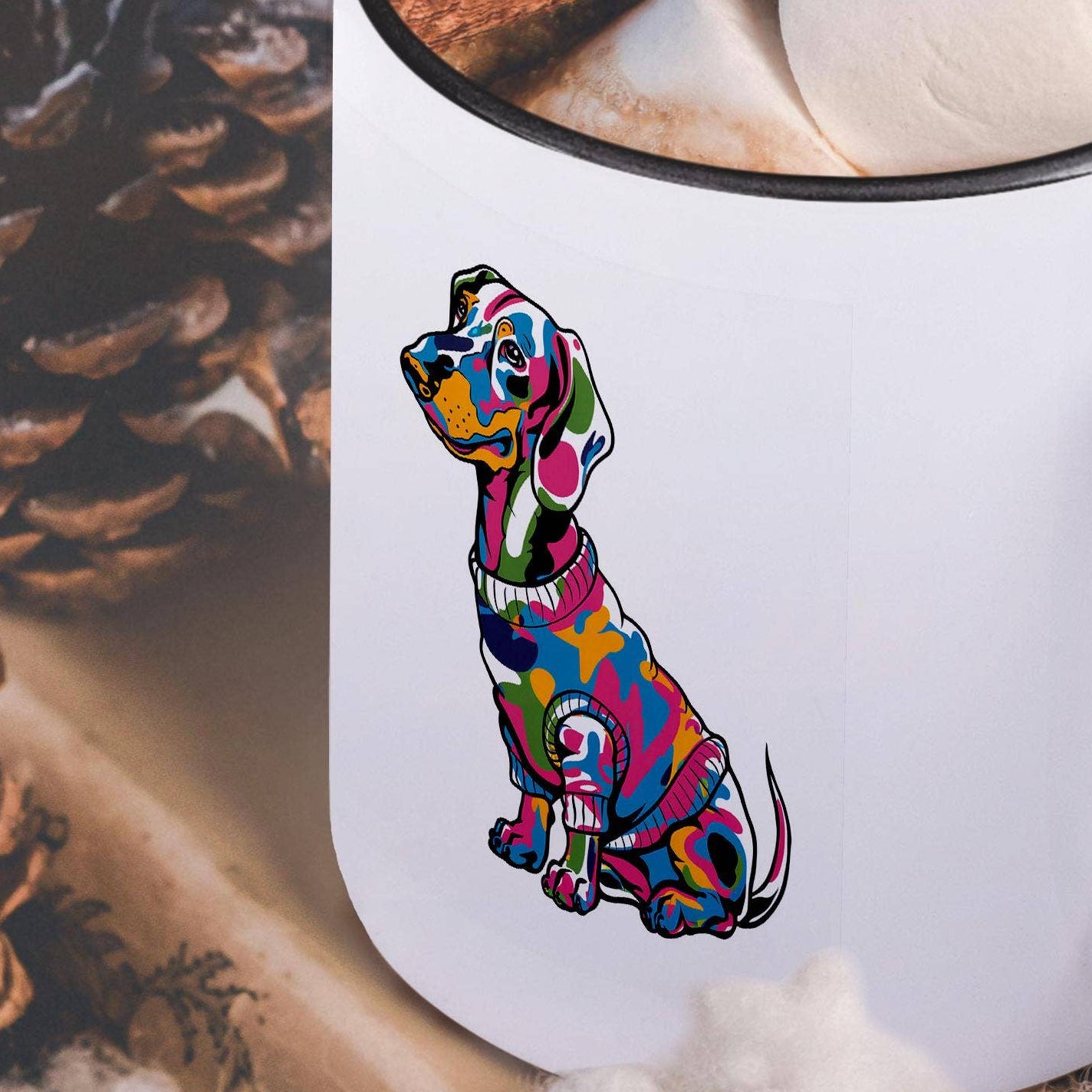 White mug with a colorful dog illustration, filled with hot chocolate and garnished with marshmallows and a cinnamon stick, on a wooden surface with pine cones and cotton.